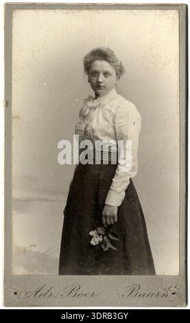 Questo ritratto in studio di Adriaan Boer mostra una giovane donna a Baarn, Paesi Bassi, tra il 1897 e il 1912. Indossa una lunga gonna scura e una camicetta dal collo alto. I suoi capelli sono modellati in un panino, e lei tiene un fiore o un ramo. La fotografia illustra la ritrattistica in studio olandese, la moda femminile, l'acconciatura, gli oggetti di scena e la rappresentazione sociale dei primi anni del XX secolo. Foto Stock
