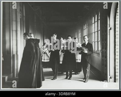 Questa fotografia del 1938 di Wiel van der Randen mostra una processione nell'abbazia benedettina di Sant'Andries vicino a Bruges, in Belgio, dopo una cerimonia di giuramento. I giovani monaci tornano dalla sala capitolare, seguiti dai membri della famiglia che portano i mantelli del coro. L'immagine mette in evidenza i rituali monastici, le processioni cerimoniali, l'abbigliamento, il coinvolgimento familiare e la sequenza strutturata della vita religiosa benedettina nel Belgio dell'inizio del XX secolo. Foto Stock