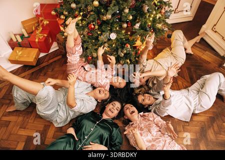 Le giovani donne in pigiama stanno ridendo e sdraiate sul pavimento nel soggiorno vicino all'albero di Natale decorato. Attrac Foto Stock