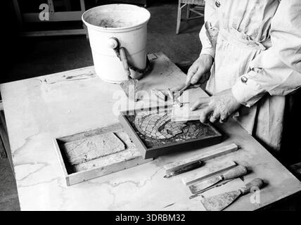 La fotografia raffigura Antoon Molkenboer a Haarlem, Paesi Bassi, 1943, che lavora su un piccolo mosaico di una Madonna. Un secchio con una cazzuola e vari attrezzi sono sul tavolo accanto al mosaico. L'immagine illustra la costruzione di mosaici, l'uso di utensili, l'allestimento dello spazio di lavoro, il processo artistico, l'arte visiva olandese della metà del XX secolo e l'artigianato dettagliato coinvolto nei mosaici a tema religioso. Foto Stock
