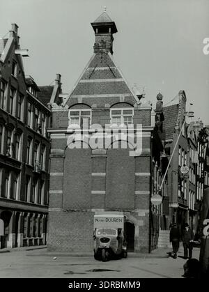 Piccolo camion a tre ruote appartenente a N. van Duuren presso il Korenmetershuisje (Persmuseum) di Amsterdam, fotografato nel 1943. Il veicolo è dotato di una ruota anteriore singola, due ruote posteriori, vano di carico chiuso, targa olandese, piantone dello sterzo, motore di piccole dimensioni e design per il trasporto di servizi di pubblica utilità della metà del xx secolo. La fotografia documenta il design dei veicoli, la logistica urbana, l'applicazione industriale e il trasporto commerciale olandese durante la guerra. Foto Stock