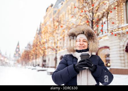 Una giovane donna con abiti casual invernali tiene una tazza di caffè e sorride all'aperto. Una donna che ride con una giacca calda e un cappello è drin Foto Stock