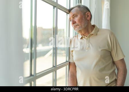 Triste stanco solo deluso anziano a casa da solo. Nonno infelice che guarda tristemente fuori dalla finestra che prova grie Foto Stock