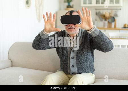 Uomo anziano entusiasta che indossa a casa occhiali VR metaversi con realtà virtuale. Nonno che tocca l'aria durante la realtà virtuale Foto Stock