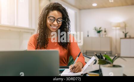 Una donna scrive in un notebook mentre è seduta alla scrivania con un notebook in un ambiente di lavoro moderno Foto Stock