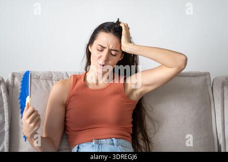 Donna stressata stanca che soffre di caldo estivo a casa, seduta sul divano che ondeggia un ventilatore di carta. Foto Stock