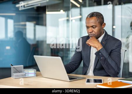 Uomo professionista che pensa profondamente e si concentra sul proprio lavoro, seduto alla scrivania con un laptop e uno smartphone, riflettendo l'attenzione concentrata in un ambiente aziendale Foto Stock