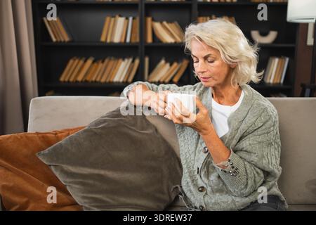 Buon mattino autunno inverno atmosfera autunnale. Confortevole anziana anziana rilassata che beve bevande calde tè e caffè nel divano di casa. Foto Stock