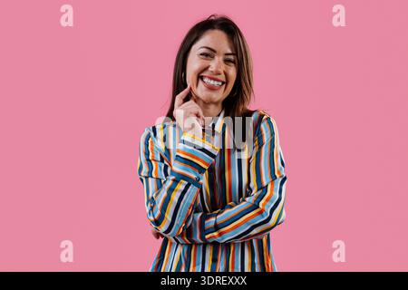 Giovane donna felice che ride e posa con una camicia colorata a righe su uno sfondo rosa brillante, esprimendo gioia, sicurezza ed energia vibrante Foto Stock