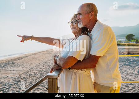 Coppia anziana amorevole in piedi su una passerella sulla spiaggia durante il tramonto, la donna che punta verso il mare mentre l'uomo la abbraccia da dietro, sh Foto Stock