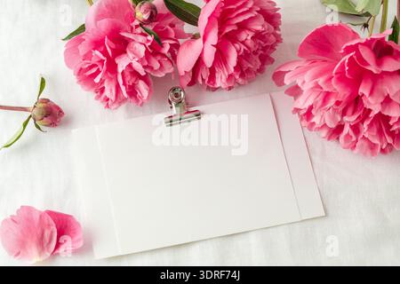 Fiori di peonia e cartolina bianca bianca su sfondo in puro lino. Tessera floreale d'invito in stile vintage. Saluto di compleanno con mockup c Foto Stock
