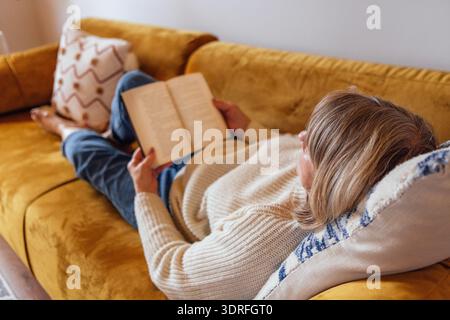 Una donna anziana in abiti casual è sdraiata sul divano e sta leggendo un romanzo. Una signora matura tiene un libro e si rilassa a casa Foto Stock