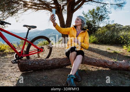 Donna anziana in abbigliamento attivo sorridente alla fotocamera mentre riposa su un tronco, prendendo una pausa dalla mountain bike in un paesaggio naturale di montagna al sole Foto Stock