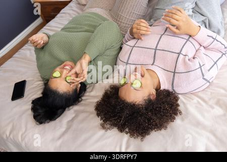Amiche asiatiche e afroamericane sdraiate a letto con maglioni in maglia con fette di cetriolo Foto Stock