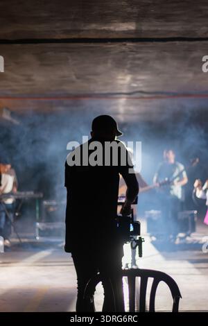 Un uomo sta di fronte a un gruppo di musicisti, a filmarli. La scena è scarsamente illuminata, con fumo sullo sfondo. La scena è misteriosa e un po' om Foto Stock