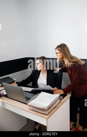Due donne lavorano insieme in un ufficio, discutendo attività e rivedendo documenti a una scrivania con un computer. Foto Stock