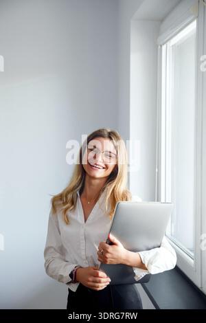 Una donna sta in un ufficio, sorride mentre tiene un notebook vicino al petto. Una luce intensa entra attraverso la finestra. Foto Stock