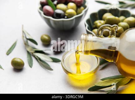 Versare olio d'oliva vergine in una piccola ciotola con olive e rami d'oliva Foto Stock