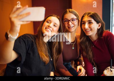 Tre ragazze carine sorridono e fanno selfie. Amici spensierati in abiti casual sono in giro in un bar. Studenti felici ar Foto Stock