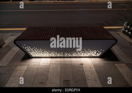 Moderna panca illuminata con lastre di legno sul marciapiede della città di notte, luci accese sotto la struttura, scena urbana Foto Stock
