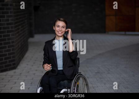 Donna d'affari sorridente in sedia a rotelle che usa uno smartphone all'esterno Foto Stock