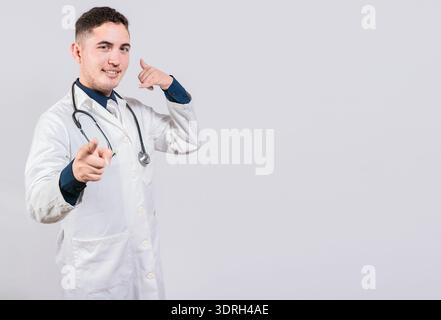Bel medico che fa il gesto di chiamata isolato. Medico sorridente che fa un gesto di chiamata e punta verso la fotocamera. Foto Stock
