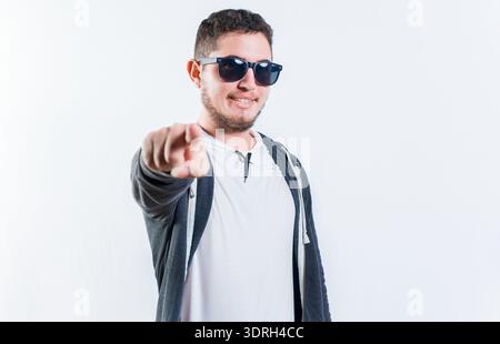 Bel ragazzo con gli occhiali da sole sorridente e che punta alla telecamera isolata Foto Stock