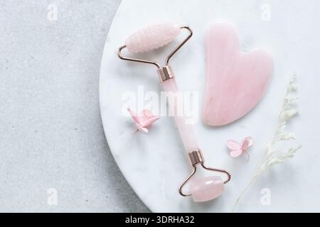 Rullo per viso in pietra e raschietto gua Sha per massaggio viso di bellezza. Concetto anti-invecchiamento della pelle. Piatto su marmo bianco Foto Stock