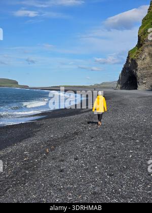 Un viaggiatore solitario con un mantello giallo brillante cammina lungo le sabbie vulcaniche nere della spiaggia di Reynisfjara, circondato da scogliere spettacolari Foto Stock
