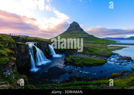 La montagna di Kirkjufell si innalza drammaticamente su una vegetazione lussureggiante e cascate sotto un cielo vibrante in Islanda Foto Stock
