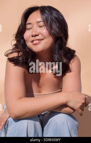 La donna asiatica con lunghi capelli scuri sorride e si siede comodamente su un morbido sfondo beige, mostrando un rilassato e gioia Foto Stock