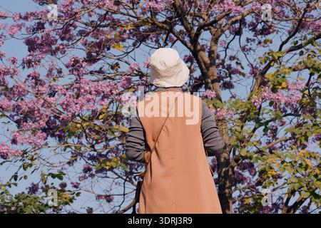 Donna che indossa un cappotto beige e un cappello bianco in piedi sotto un albero viola fiorito in una soleggiata scena all'aperto Foto Stock