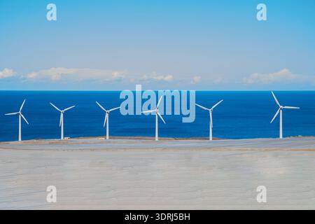 Turbine eoliche sul paesaggio costiero con cielo azzurro cristallino e oceano sullo sfondo che mettono in mostra energia rinnovabile Foto Stock