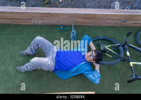 Uomo steso su erba artificiale, che si prende una pausa dal ciclismo e dal giardinaggio Foto Stock