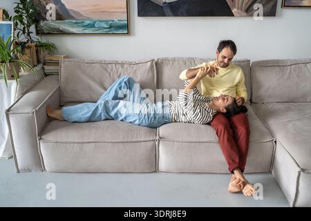 Felice coppia di sposi uomo e donna che si rilassano a casa sul divano insieme, divertendosi sorridendo e scherzando. Foto Stock
