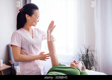 La terapista femminile in scrub rosa sta fornendo terapia fisica a una donna sorridente in camicia verde, dimostrando una guarigione di sostegno Foto Stock
