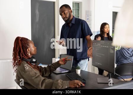 Gli sviluppatori maschi e femmine di colore esaminano il codice sorgente HTML sul monitor del pc e sul laptop durante la riunione. I colleghi comunicano le idee in modo chiaro mentre realizzano soluzioni digitali negli uffici moderni. Foto Stock