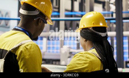 Membri del personale IT che sovrintende alle linee di produzione di pannelli solari e alle apparecchiature. Colleghi che eseguono brainstorming, manutenzione di sistemi di assemblaggio fotovoltaici in fabbrica 4,0 automatizzata, telecamera B. Foto Stock