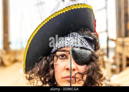 Donna vestita da capitano pirata che indossa un cappello tricorno, un occhiello, una bandana e un trucco, tenendo una cutlass davanti al viso, incarnando l'avventura An Foto Stock