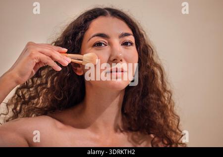 Donna con capelli ricci che applica il trucco con una spazzola, che mostra routine di bellezza e cura di sé, con illuminazione soffusa e bac neutro Foto Stock