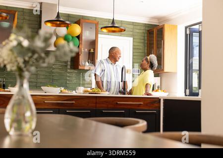 Coppia senior che si diverte a conversare in una cucina moderna con palloncini festivi. Anziani, festeggiamenti, famiglia, stile di vita, insieme Foto Stock
