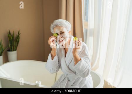 Matura donna anziana adulta con nutriente anti età maschera crema viso tenendo fette di cetriolo. Donna anziana con maschera bianca creme su S. Foto Stock