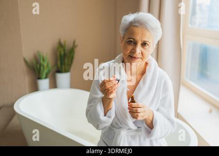 Donna che applica olio cosmetico a gocce naturali sulla pelle del viso. Donna anziana adulta matura che applica siero nutriente anti età con la pipetta Foto Stock