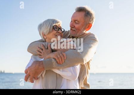 Coppie anziane che si abbracciano godendosi le attività ricreative all'aperto a piedi sulla spiaggia. Vecchio marito moglie che abbraccia con tenerezza amore godere Foto Stock