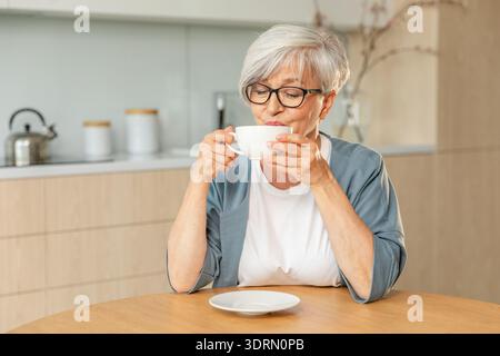 Buon giorno. Comfort rilassato anziano anziano anziano anziano donna nonna che beve bevande calde tè e caffè in cucina a casa. Maturo W Foto Stock