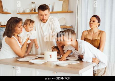 Famiglia che festeggia il primo compleanno di un bambino con una torta su un tavolo da cucina, circondata da espressioni gioiose e atmosfere festose Foto Stock