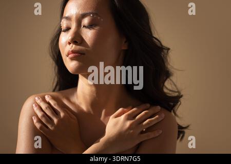Donna asiatica con lunghi capelli scuri, che si abbraccia delicatamente, sfoggia bellezza naturale e amore per se stessa, illuminata da luce soffusa ag Foto Stock