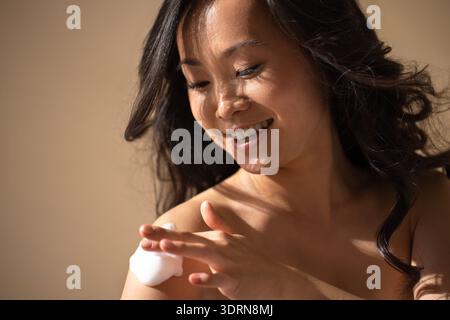 Donna asiatica che applica crema per la pelle alle spalle, sorride dolcemente, con un'illuminazione soffusa che mette in risalto la sua bellezza naturale e crea Foto Stock
