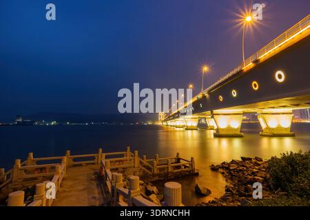 Le calde luci dorate di un ponte illuminato si riflettono magnificamente sulle acque calme sotto un cielo blu intenso al crepuscolo. Foto Stock