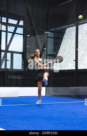 Atleta in abito nero con racchetta oscillante in padel che colpisce la palla da tennis su un campo blu con pareti in vetro. Sport, fitness, competitio Foto Stock